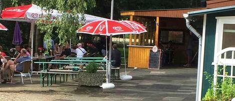 An outdoor dining area with tables and chairs, umbrellas, and a small building in the background.