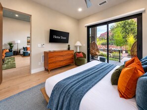 Een moderne slaapkamer met een groot bed, een televisie en een schuifdeur naar een balkon met uitzicht op groen en een gebouw.