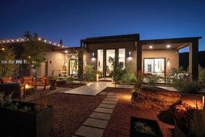 Een modern huis met een overdekte patio, buitenmeubels en een verlichte tuin 's nachts.