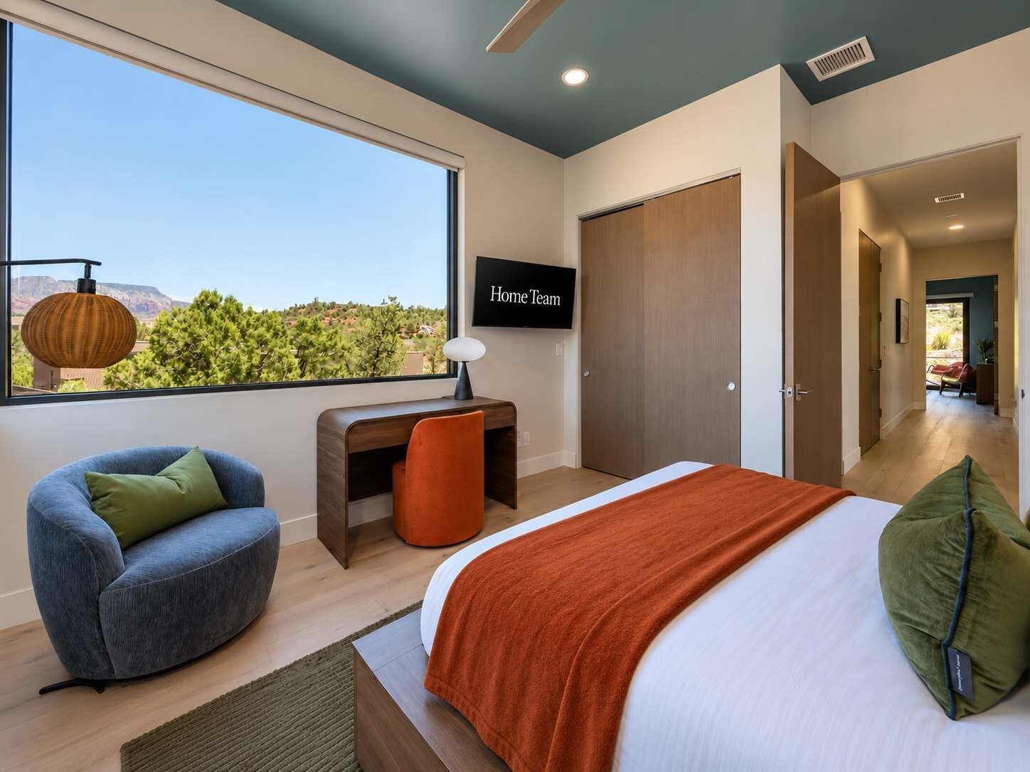Un dormitorio moderno con un ventanal grande, una cama con colcha naranja, un escritorio y una silla.