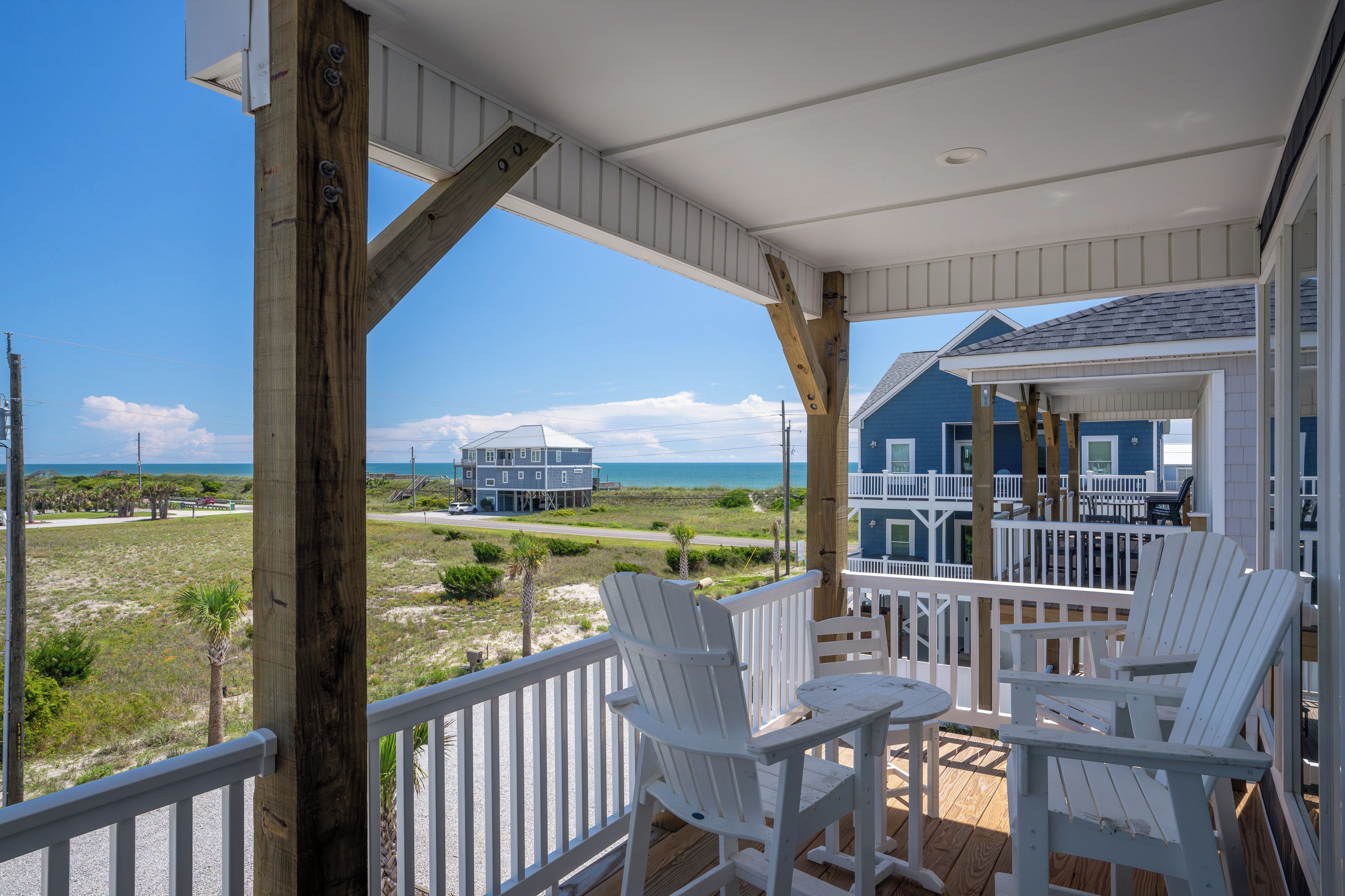 Eine Holzterrasse mit weißen Stühlen und einem Tisch, die auf einen Strand und ein Haus mit blauer Fassade blickt.