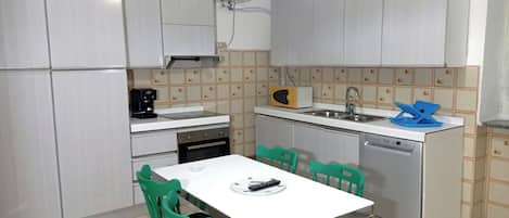 A compact kitchen with a white countertop, a coffee maker, a microwave, a sink, and a dishwasher. There are green chairs around a small dining table.