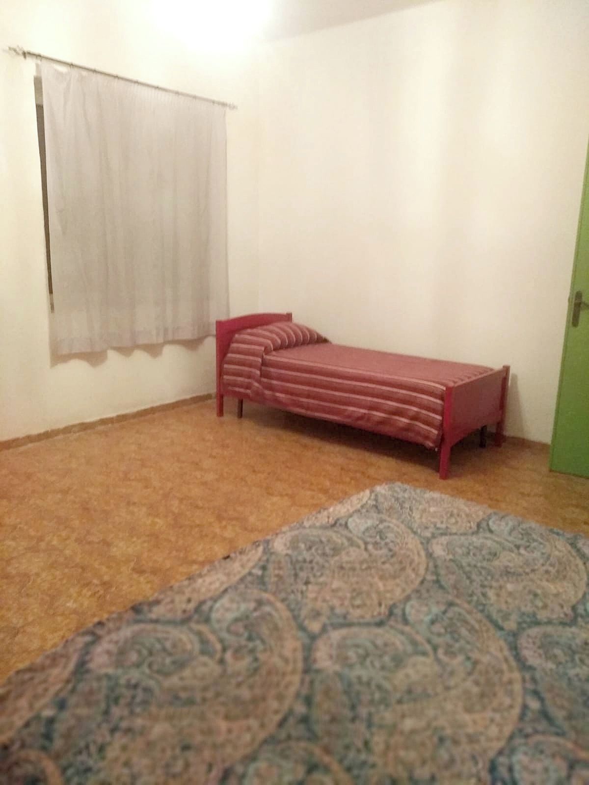 Ein kleines Zimmer mit einem Einzelbett, einem gemusterten Teppich, einem Fenster mit Vorhang und einer grünen Tür.