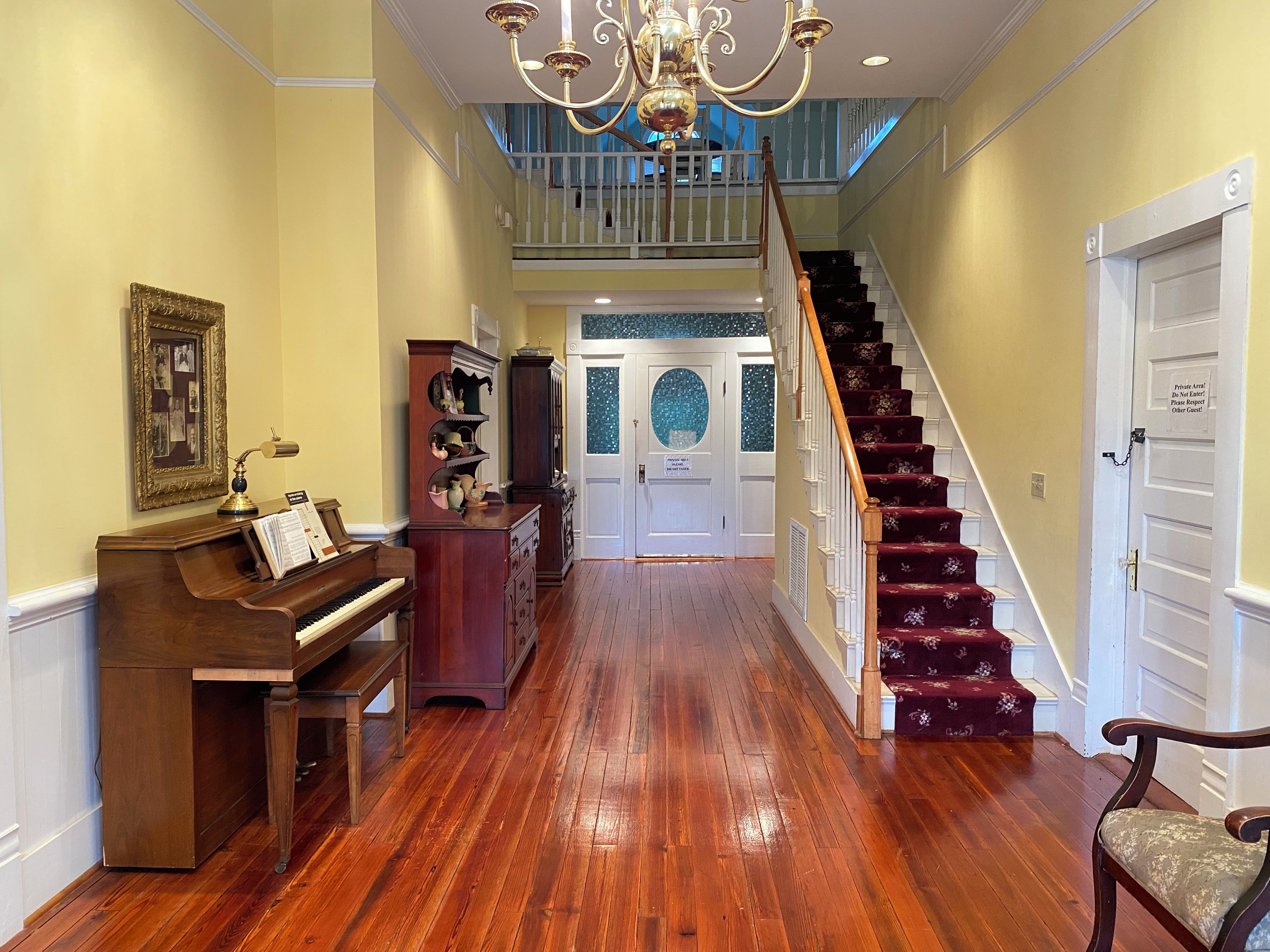 A grand hallway with a piano, a wooden cabinet, a staircase with red carpet, and a chandelier.