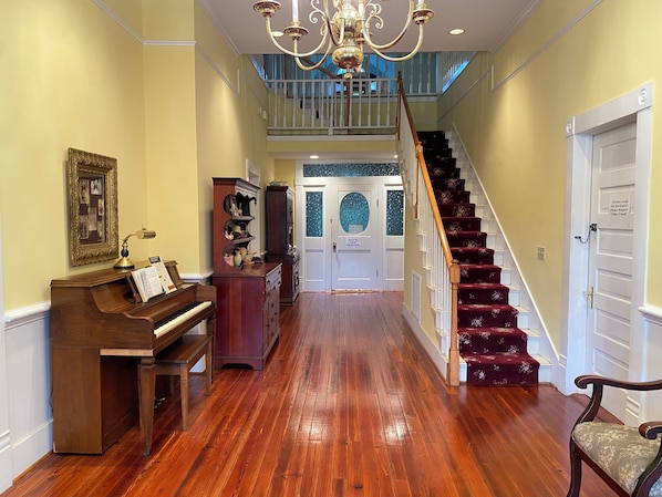 A grand hallway with a piano, a wooden cabinet, a staircase with red carpet, and a chandelier.