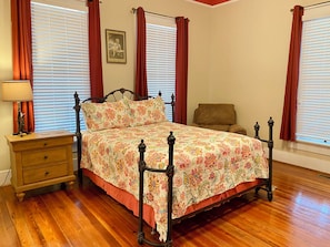 A bedroom with a wooden floor, a bed with a floral bedspread, a nightstand with a lamp, and a chair.
