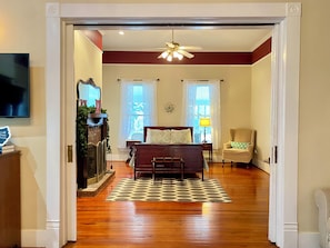 A bedroom with a wooden floor, a bed with a patterned bedspread, a chair, a nightstand, a lamp, and a ceiling fan. - This Old House (Goshen)
