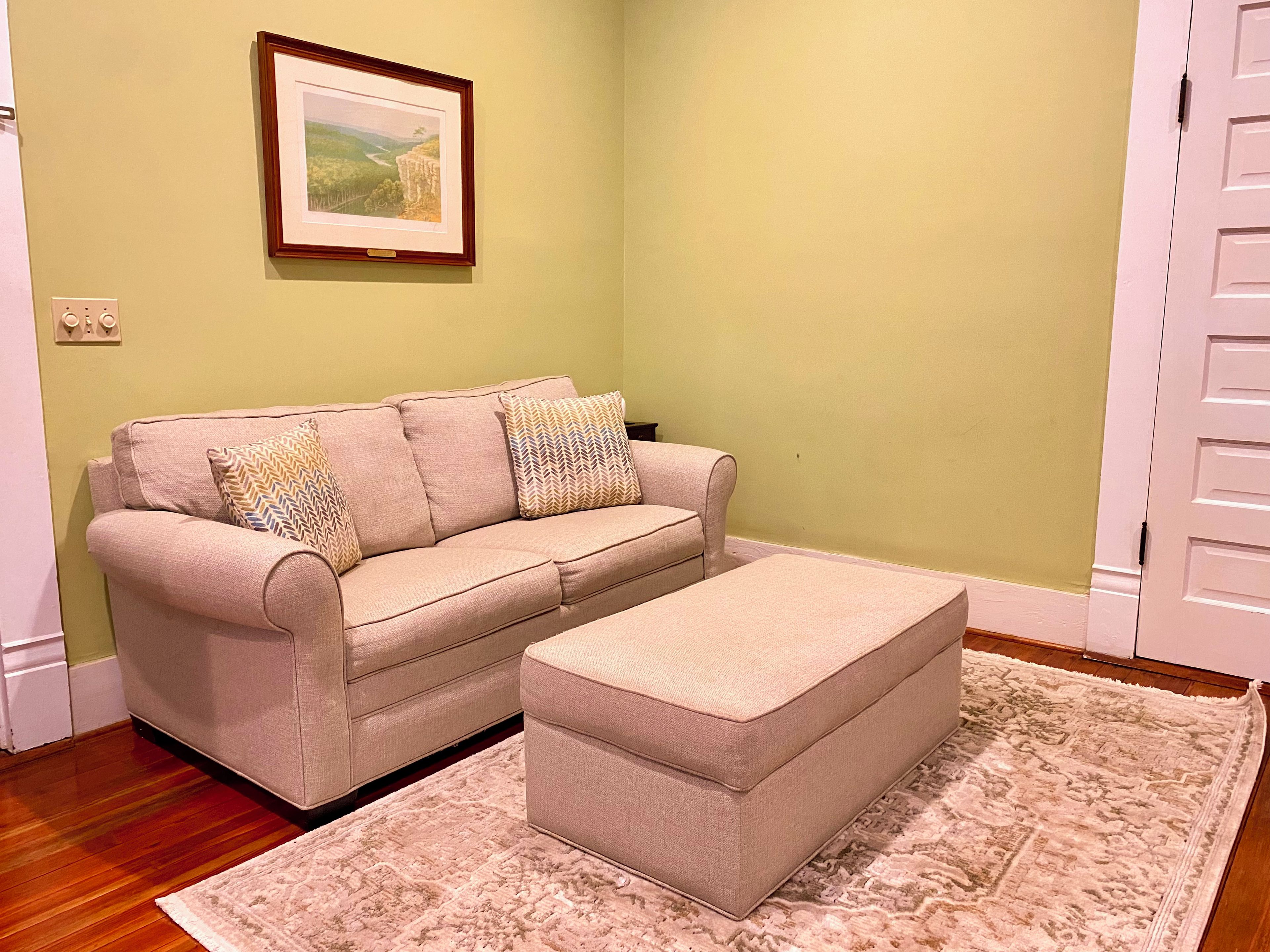Ein Wohnzimmer mit einem beigefarbenen Sofa, passendem Hocker und einem gemusterten Teppich.