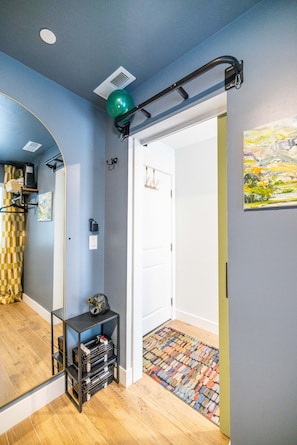 Un pasillo con un espejo, una estantería negra con objetos y una puerta que da a otra habitación, además de una alfombra colorida.