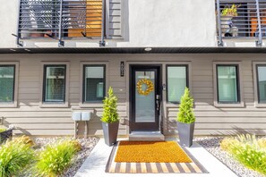 Un bâtiment moderne à deux étages, doté d’une porte d’entrée, de deux plantes en pot, d’un paillasson et d’un petit jardin agrémenté de diverses plantes.