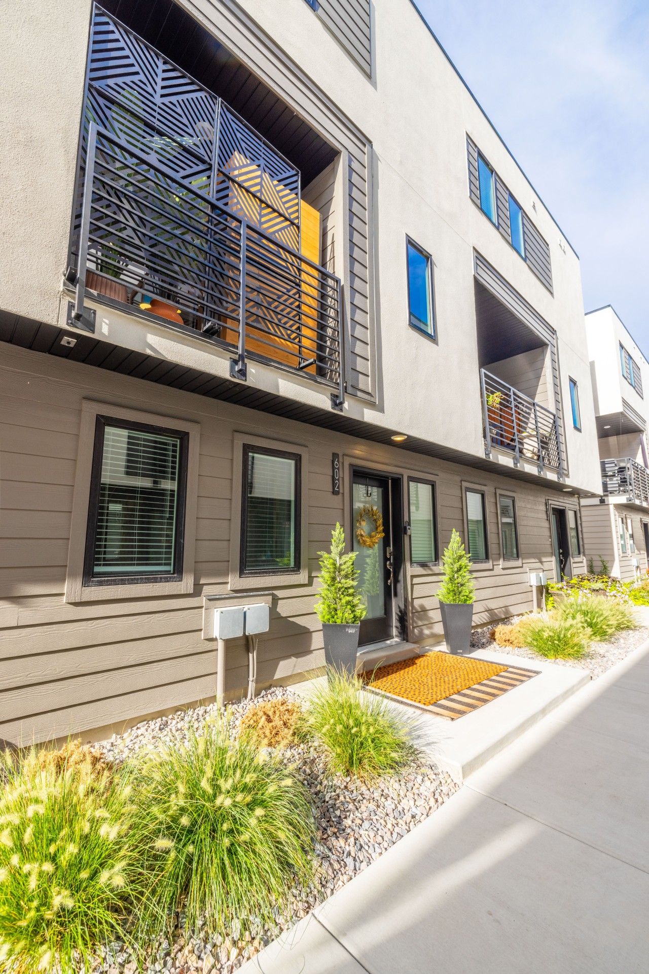 A modern multi-story residential building with balconies, a front door with a wreath, and a landscaped garden with various plants.