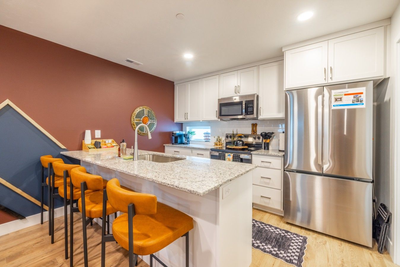 A modern kitchen with white cabinets, stainless steel appliances, a granite countertop, and a bar area with orange chairs.