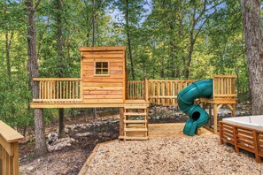 A wooden play structure with a slide and a small cabin-like building, surrounded by trees. - 7BR w Treehouse, Hot Tub, Sauna, Theater. Pets OK! (McGaheysville)