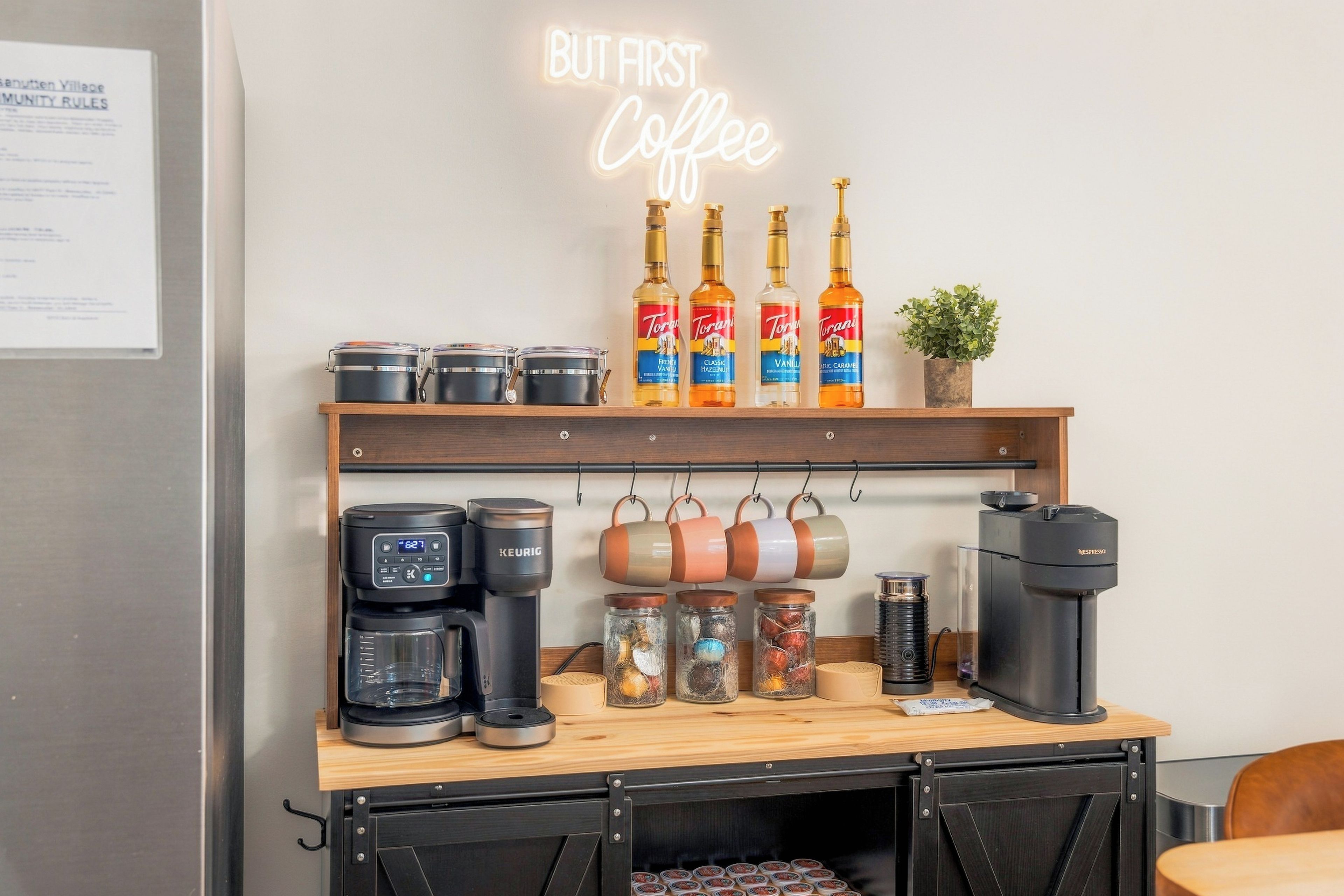 Eine Kaffeestation mit Kaffeemaschine, Kaffeebohnen in Gläsern, Torani-Sirups und einem Neon-Schild mit der Aufschrift 'BUT FIRST Coffee'.