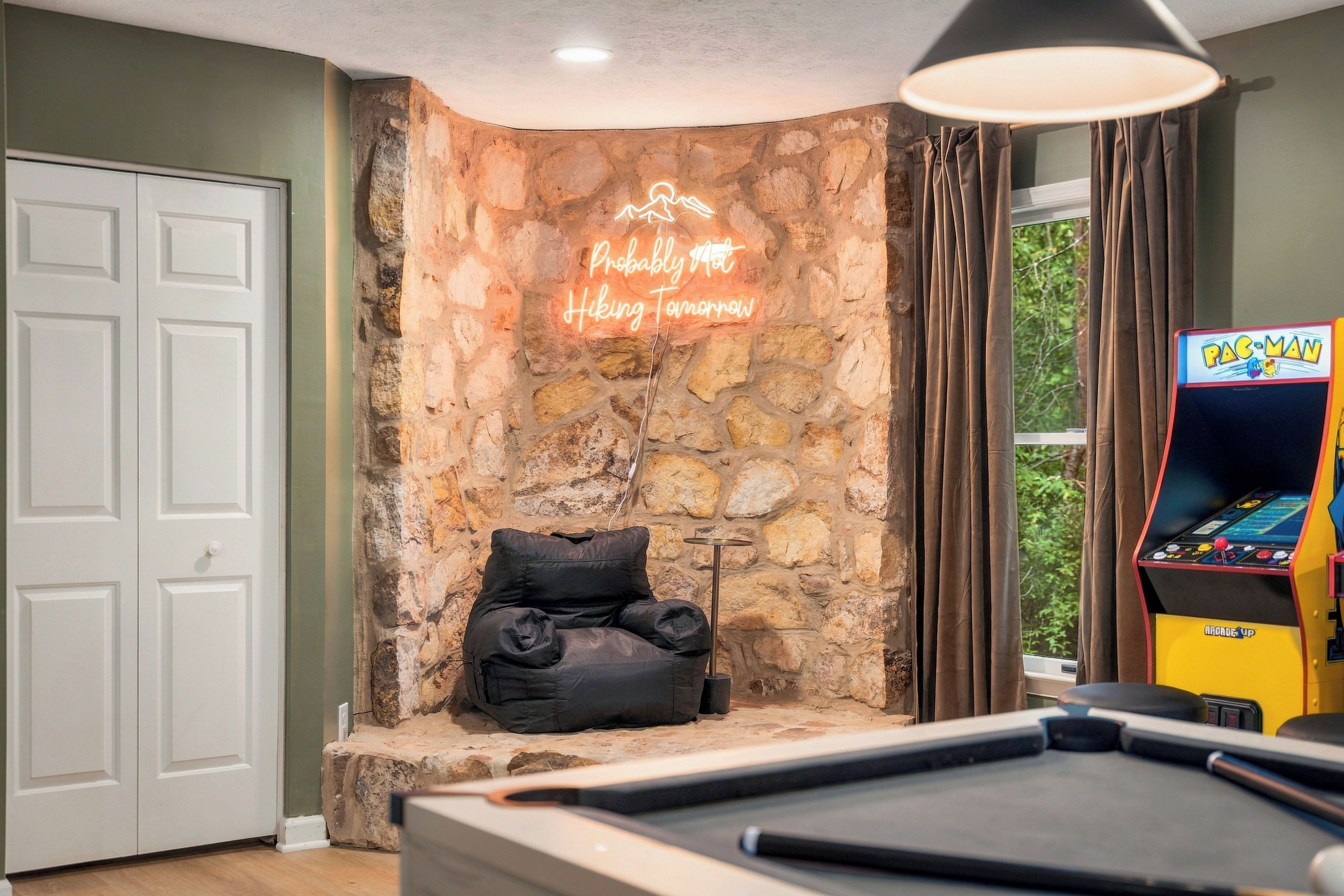 A pool table in a room with a stone wall, a neon sign, a couch, and a Pac-Man arcade machine.