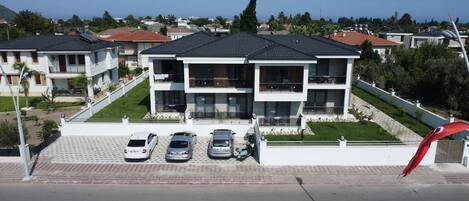 A two-story residential building with a flat roof, a balcony on the upper floor, and a driveway with three cars parked.
