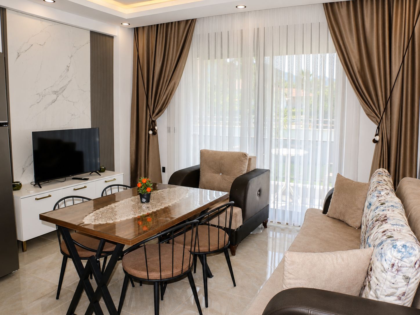 A modern living room with a flat-screen TV, a dining table with chairs, a sofa, and a large window with curtains.