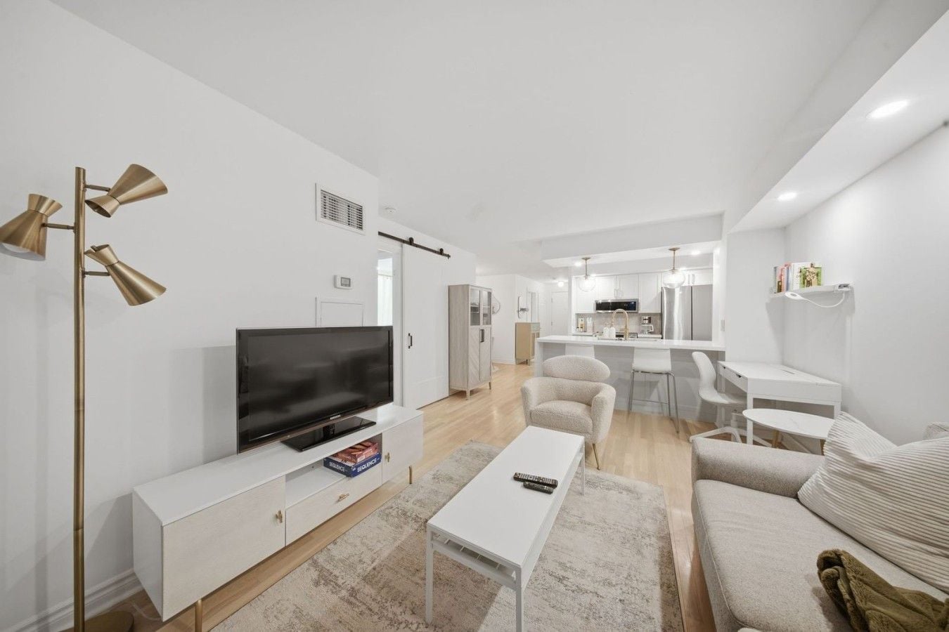 A modern living room with a flat-screen TV, white furniture, and a kitchen in the background.