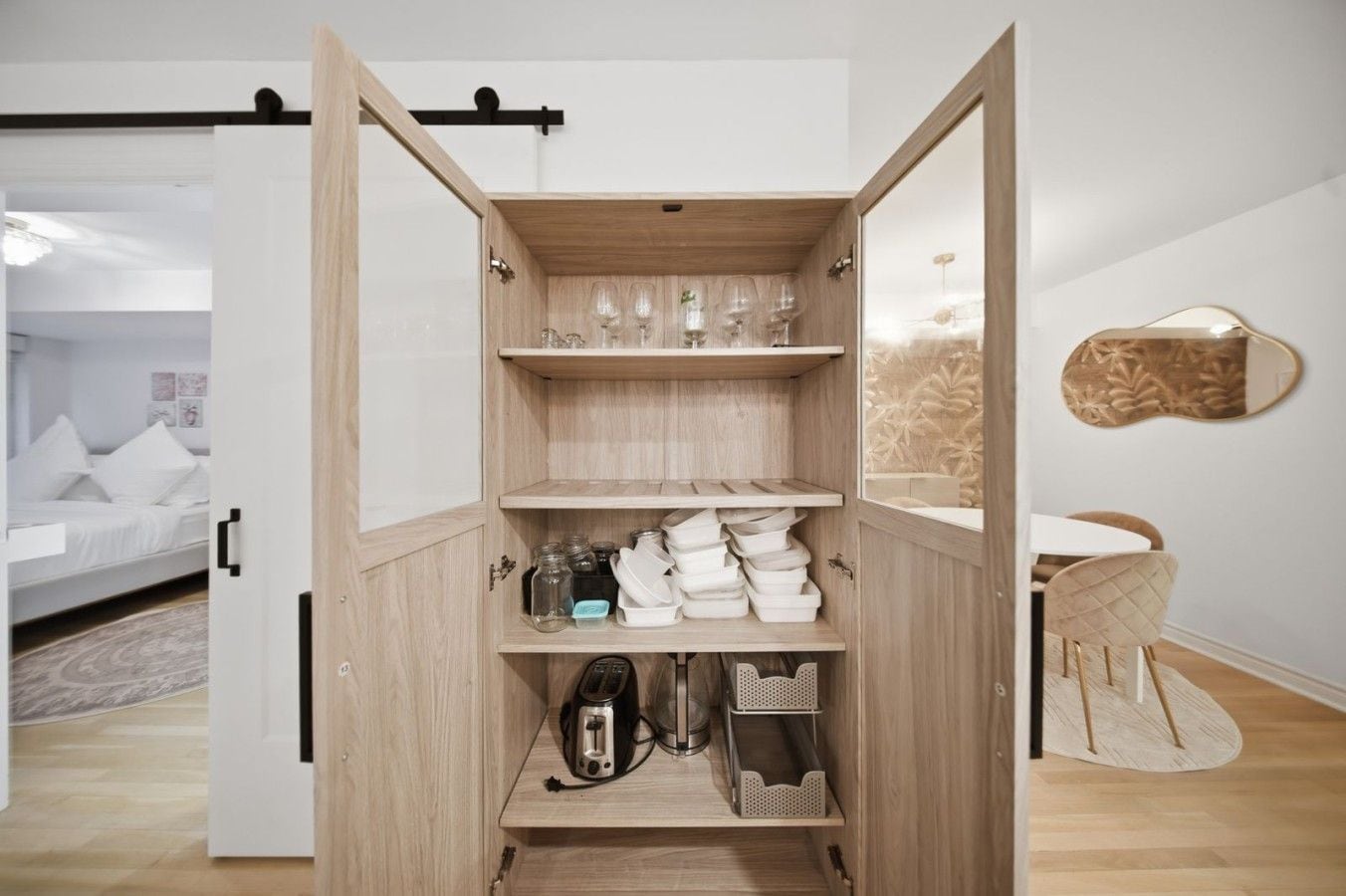 A modern, open shelving unit with kitchenware and glassware, a bed with white bedding, and a minimalist bathroom with a round mirror.