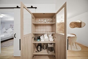 Ein modernes, offenes Regalsystem mit Geschirr und Gläsern, ein Bett mit weißer Bettwäsche und ein minimalistisches Badezimmer mit einem runden Spiegel.