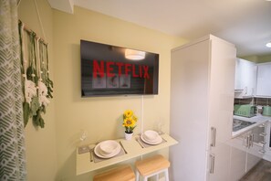 A modern kitchen with a mounted television displaying 'NETFLIX', a dining area with a vase of sunflowers, and a kitchen counter with a sink and appliances.