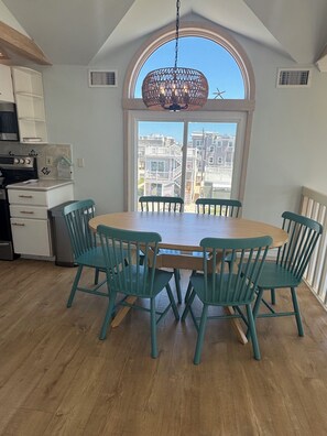 A dining area with a round table and six chairs, a kitchen with a stove and cabinets, and a large window with a view of buildings. - Bayside Beauty w 180 degree Water Views, Hot Tub, & EV Charger! (Beach Haven)