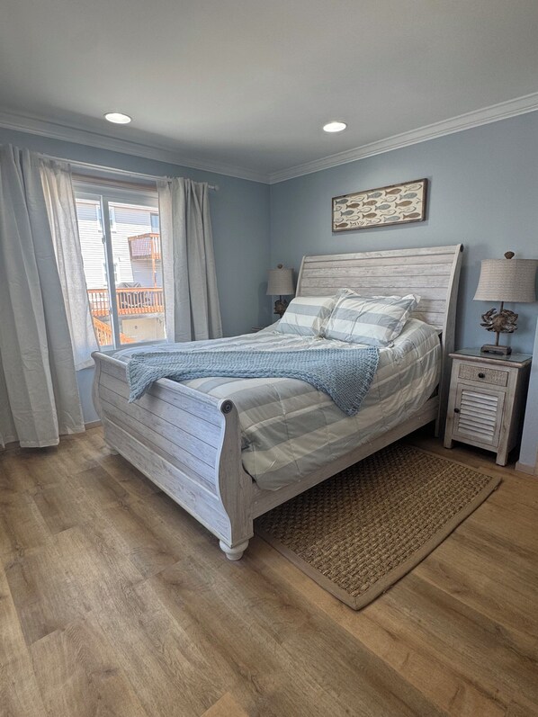 A bedroom with a bed, a nightstand, a lamp, a window with curtains, and a view of a balcony.