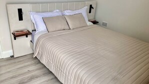 A neatly made bed with a beige quilt and pillows, a wooden nightstand with a lamp, and a wall-mounted power outlet. - Independent, Modern Annexe in Orpington (Orpington)
