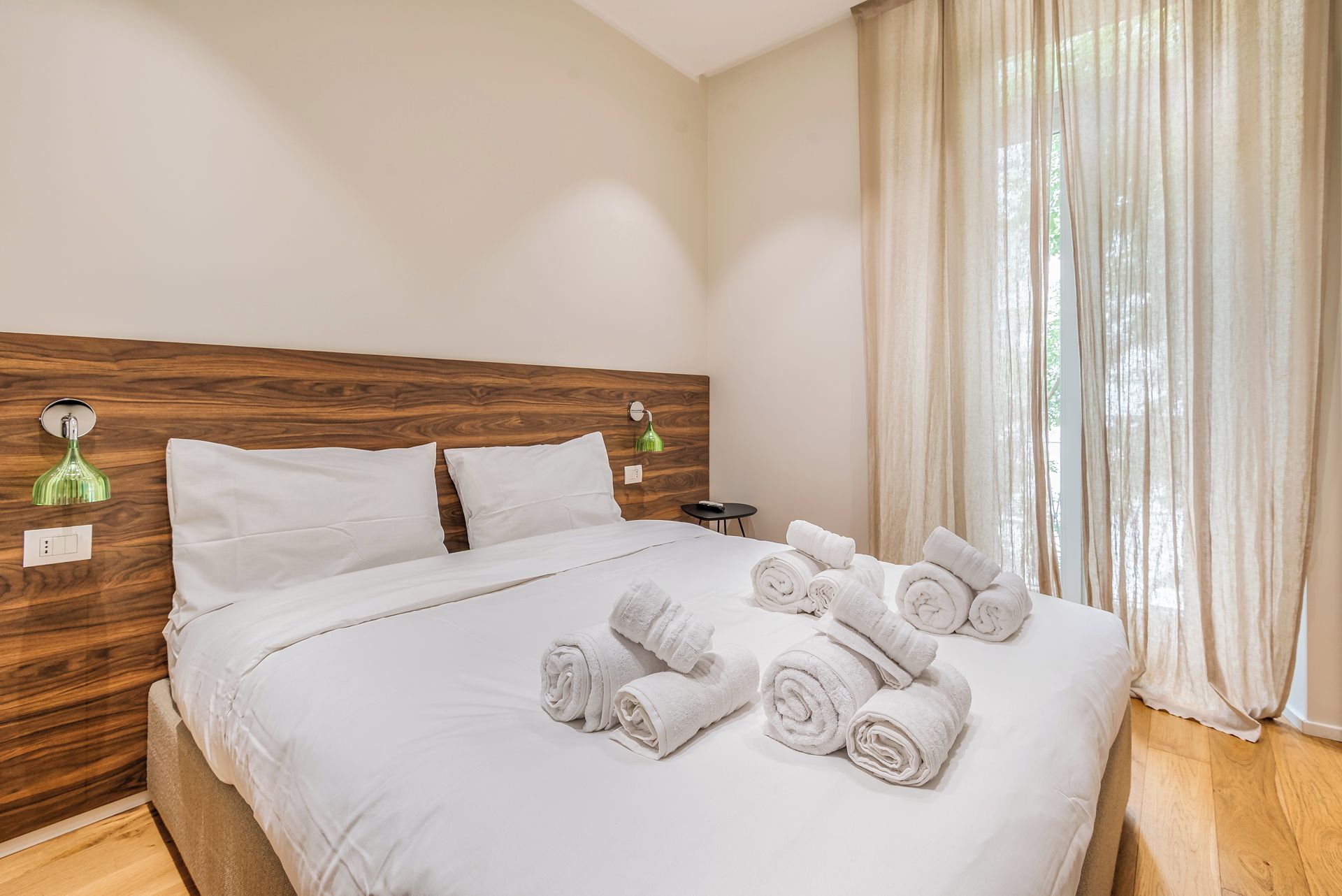 Une chambre d’hôtel avec une tête de lit en bois, un lit avec du linge de lit blanc, une lampe de chevet verte et une fenêtre avec des rideaux légers.