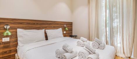 Habitación de hotel con cabecera de madera, cama con ropa blanca, lámpara de pared verde y ventana con cortinas traslúcidas.