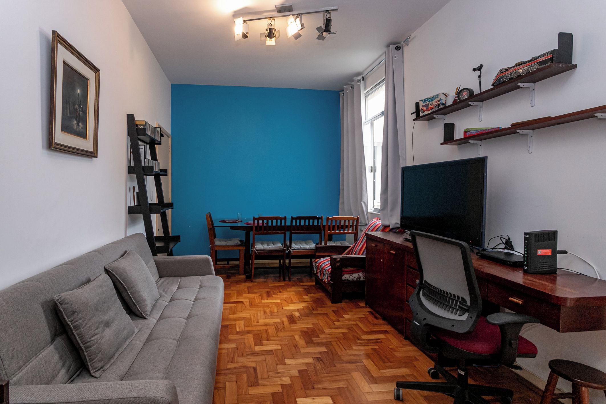 Un salon comprenant un canapé, un téléviseur, un bureau avec un ordinateur, une bibliothèque et un coin repas avec des chaises et une table.