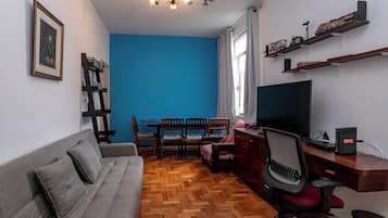 Un salon comprenant un canapé, un téléviseur, un bureau avec un ordinateur, une bibliothèque et un coin repas avec des chaises et une table.