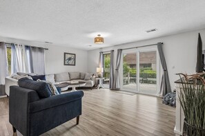 Ein großzügiges Wohnzimmer mit einer Couch, einem Couchtisch und einem Fernseher. Eine Schiebeglastür führt in einen Außenbereich.