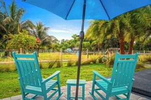 Two turquoise chairs and a small table under a blue umbrella, set on a paved area with grass and palm trees in the background. - Farm Stay, 5beds / 4.5 baths, Palm Tree, Alpaca & Llama Farm, 5 Acres (West Palm Beach)