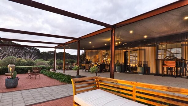 An outdoor seating area with a wooden bench, surrounded by a covered patio with a view of a building interior.