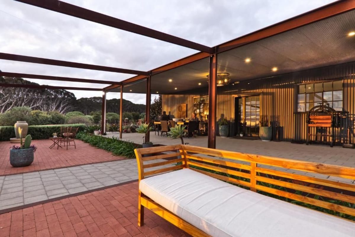 An outdoor seating area with a wooden bench, surrounded by a covered patio with a view of a building interior.