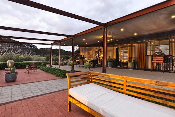 An outdoor seating area with a wooden bench, surrounded by a covered patio with a view of a building interior.