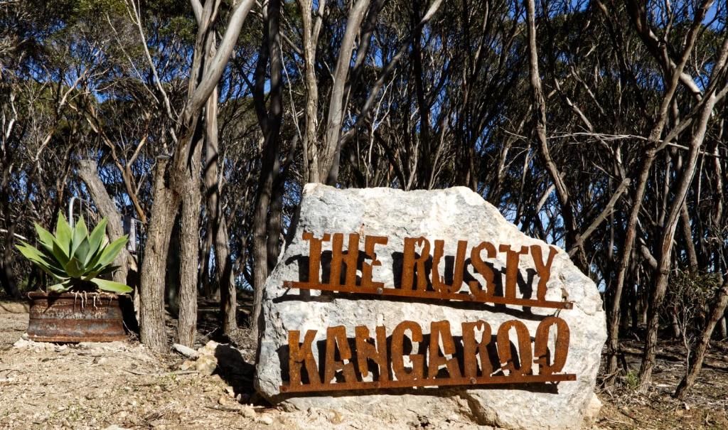 Een grote rots met de tekst 'The Rusty Kangaroo' erin gekerfd, omringd door bomen.