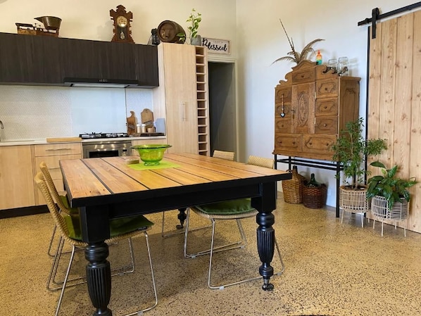 Un comedor con una mesa de madera, sillas verdes, una cocina al fondo y un mueble de madera con plantas.