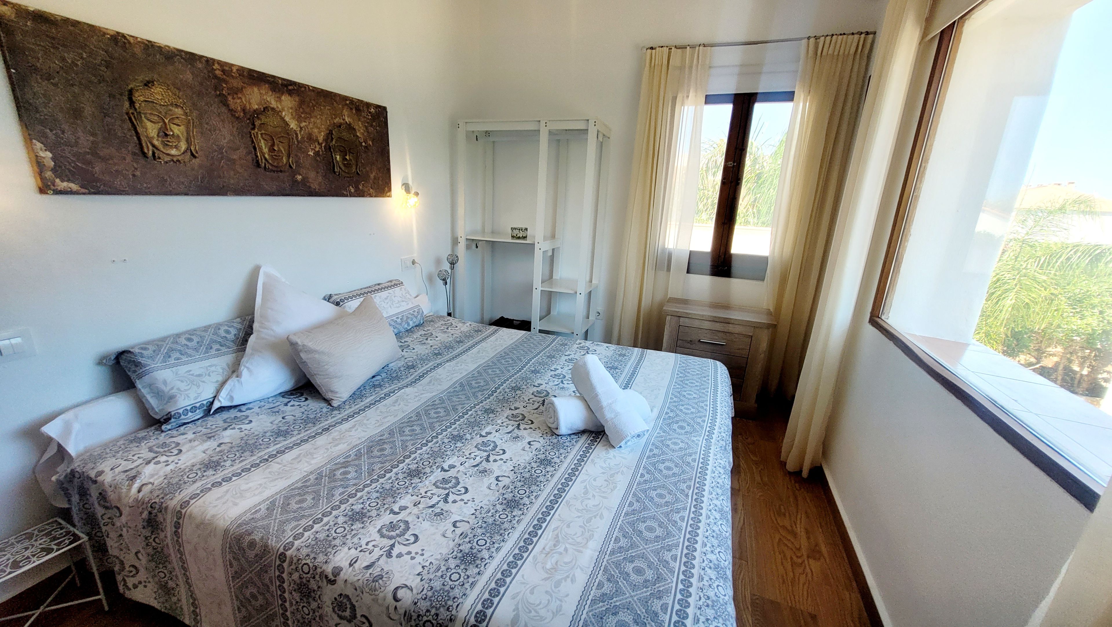 Ein Schlafzimmer mit einem Bett, Kissen, einem Nachttisch, einem Fenster mit Vorhängen und einem an der Wand hängenden Kunstwerk.