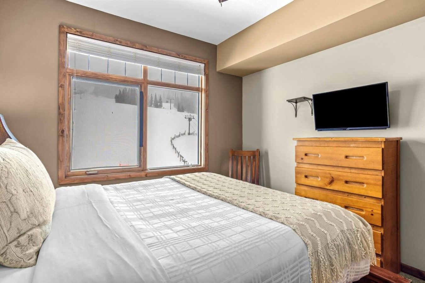 A bedroom with a bed, a wooden dresser, a television, and a window with a view of a snowy landscape.