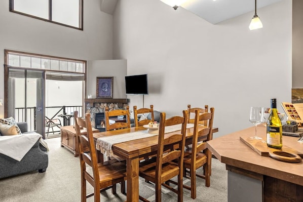 A dining area with a wooden table and chairs, a television mounted on the wall, and a bottle of wine on the table.