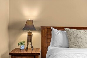 A neatly made bed with white linens and a patterned pillow, a wooden nightstand with a lamp and a small potted plant, and a plain wall.