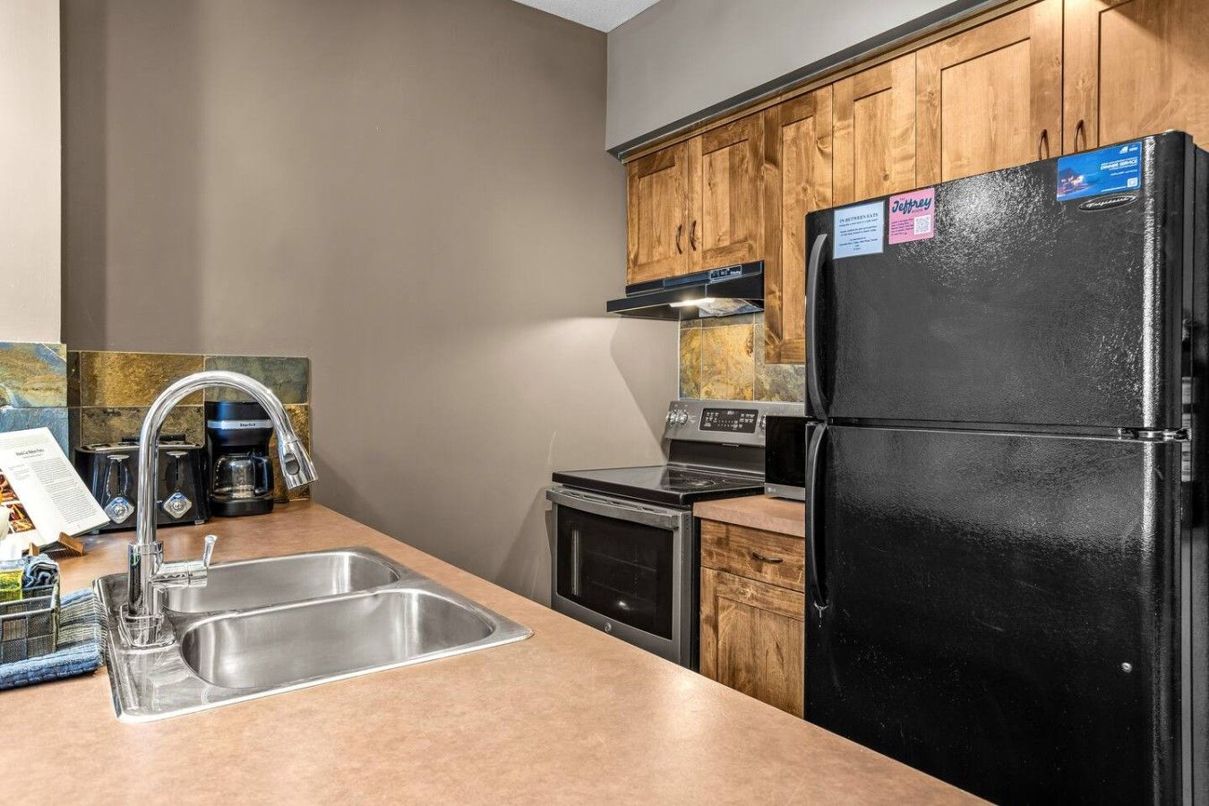 A kitchen with a stainless steel sink, a black refrigerator, wooden cabinets, a stove, and a coffee maker.