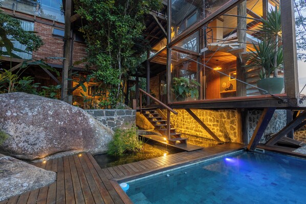 Un'area piscina moderna all'aperto con un solarium in legno, pareti in pietra e un livello superiore con vetrate e una scala.