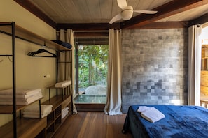 A room with a bed, a wooden shelf with towels, a ceiling fan, and a view of greenery outside. - Residencial Cambaquara 4 (Ilhabela)