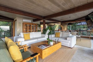 A spacious living area with a white sofa set, a wooden coffee table, and a view of the outdoors through large windows. - Residencial Cambaquara 4 (Ilhabela)