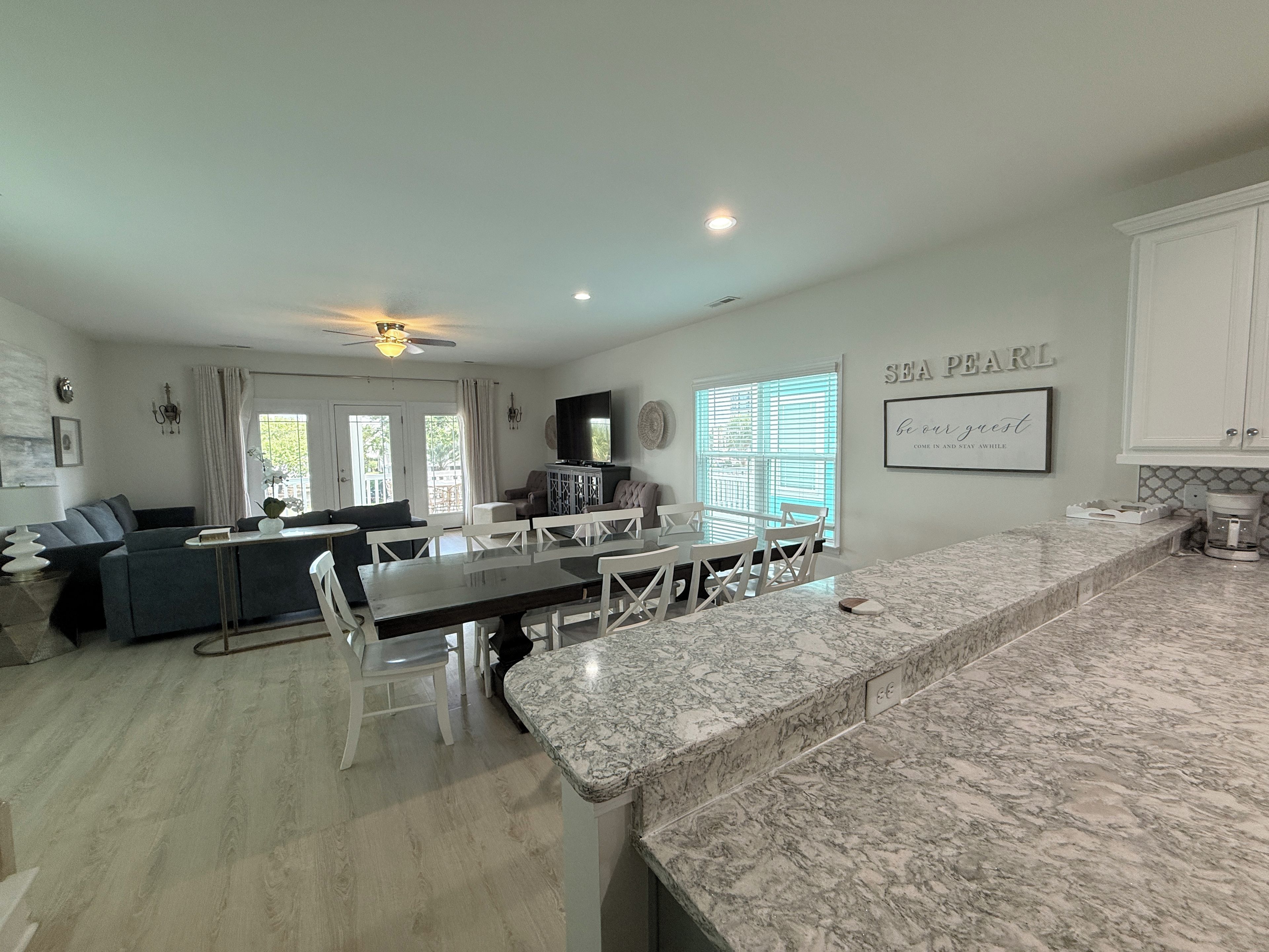 A modern kitchen with a marble countertop, a dining area with white chairs, and a living room with a sofa and a television.