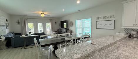 A modern kitchen with a marble countertop, a dining area with white chairs, and a living room with a sofa and a television.
