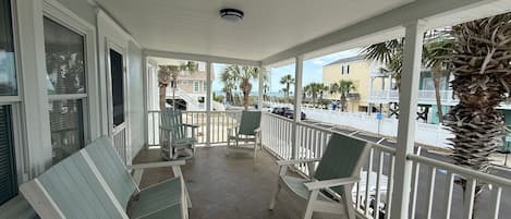 A covered outdoor area with two rocking chairs and a white railing, overlooking a beach and buildings.